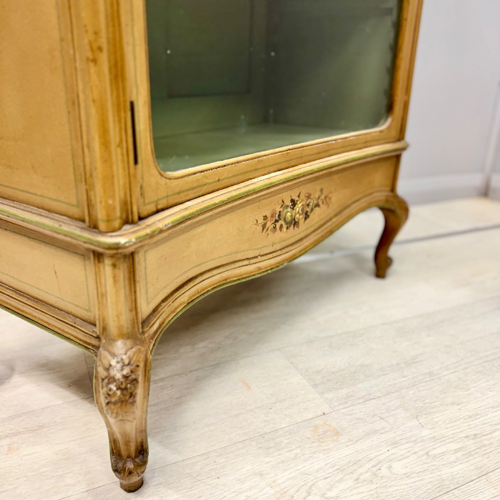 A freestanding cream painted bookcase featuring a single curved glass door, floral painted motifs, cabriole legs, and a sage green interior with adjustable shelves. View II