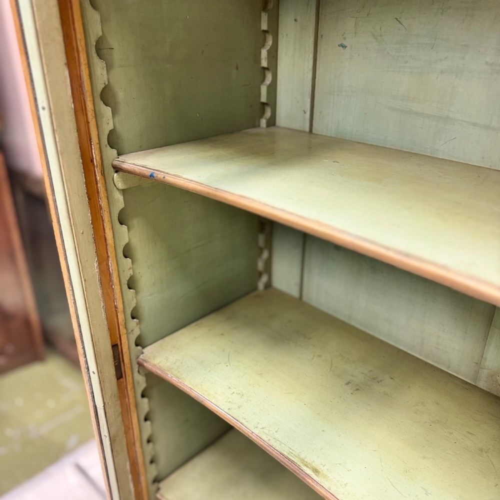 A freestanding cream painted bookcase featuring a single curved glass door, floral painted motifs, cabriole legs, and a sage green interior with adjustable shelves. View VI