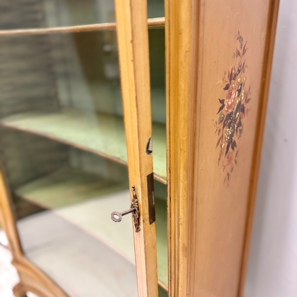 A freestanding cream painted bookcase featuring a single curved glass door, floral painted motifs, cabriole legs, and a sage green interior with adjustable shelves. View VII