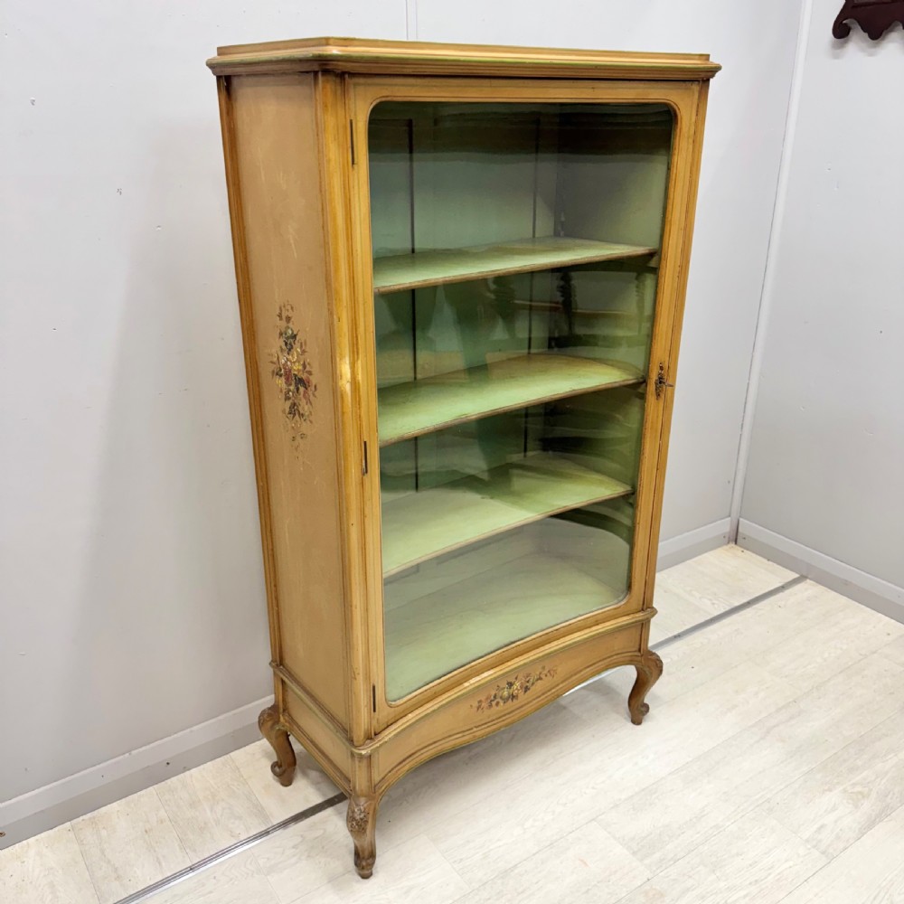 A freestanding cream painted bookcase featuring a single curved glass door, floral painted motifs, cabriole legs, and a sage green interior with adjustable shelves. View X