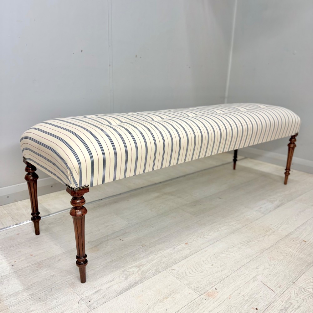 An elongated antique footstool featuring button-tufted ticking stripe upholstery and dark turned wooden legs.