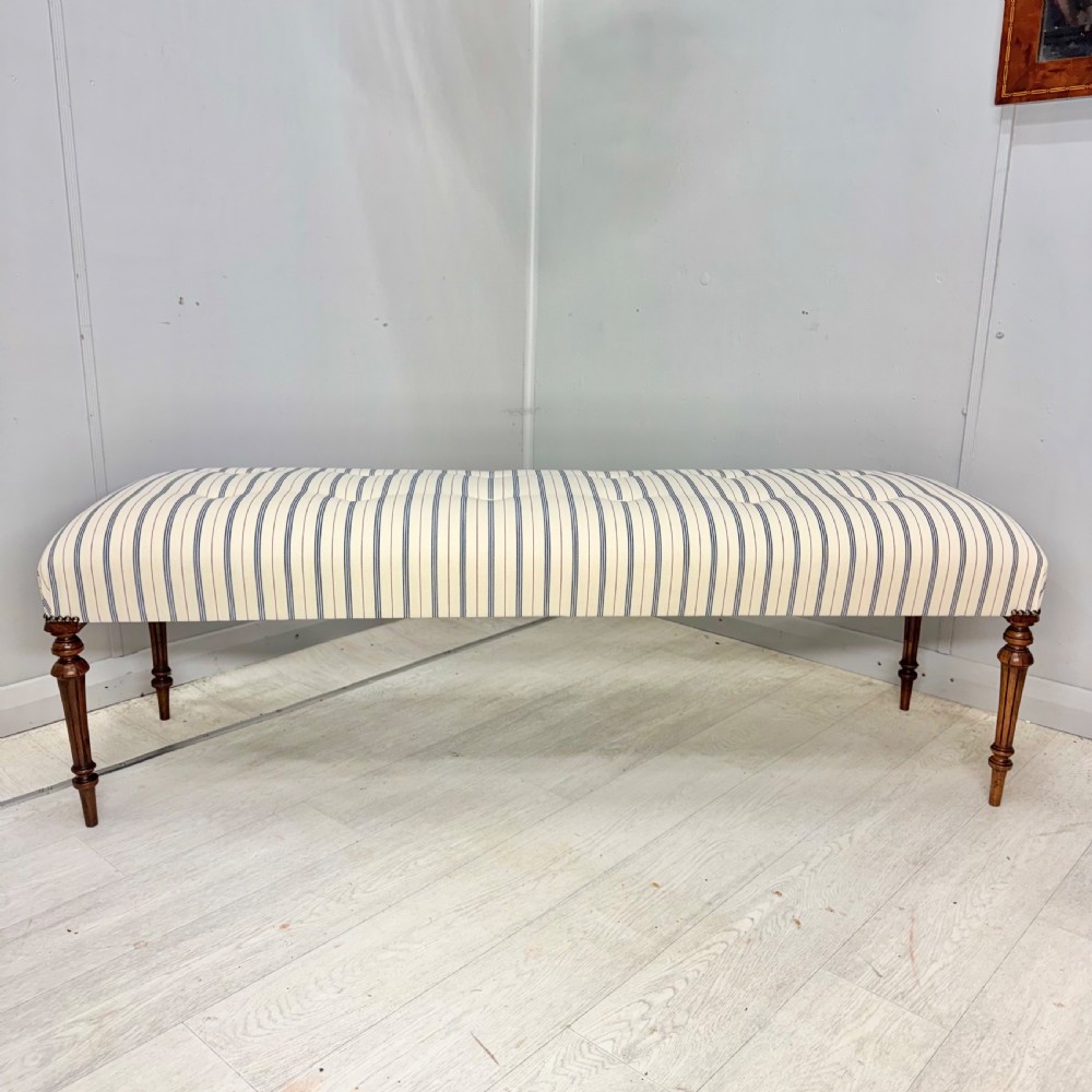 An elongated antique footstool featuring button-tufted ticking stripe upholstery and dark turned wooden legs. View V