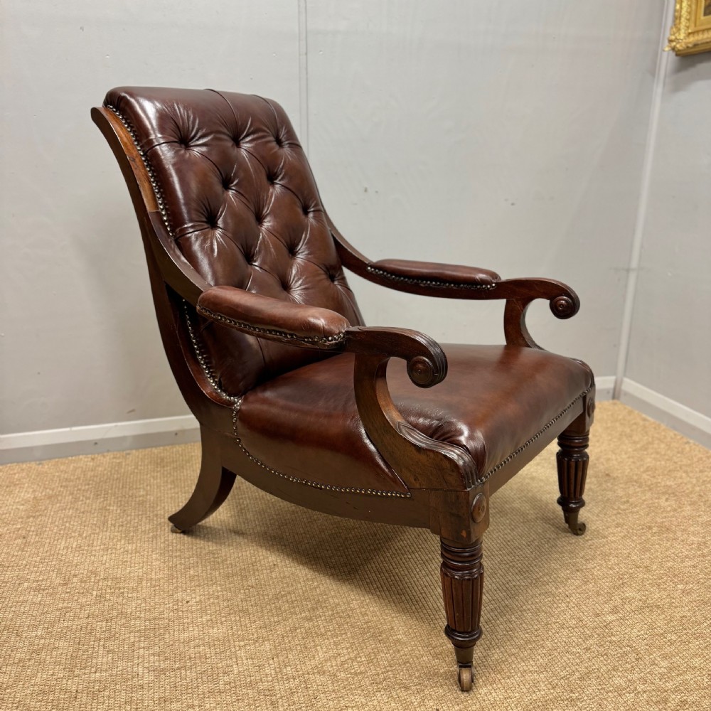 A Georgian mahogany armchair upholstered in deep brown buttoned leather with padded scroll arms, brass studded trim, and fluted front legs on castors.
