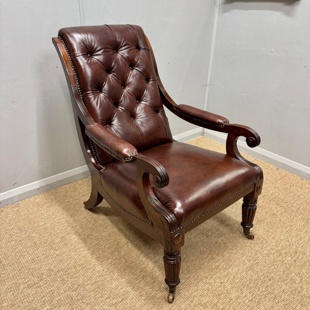 A Georgian mahogany armchair upholstered in deep brown buttoned leather with padded scroll arms, brass studded trim, and fluted front legs on castors. View II