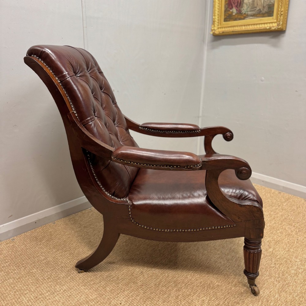 A Georgian mahogany armchair upholstered in deep brown buttoned leather with padded scroll arms, brass studded trim, and fluted front legs on castors. View V