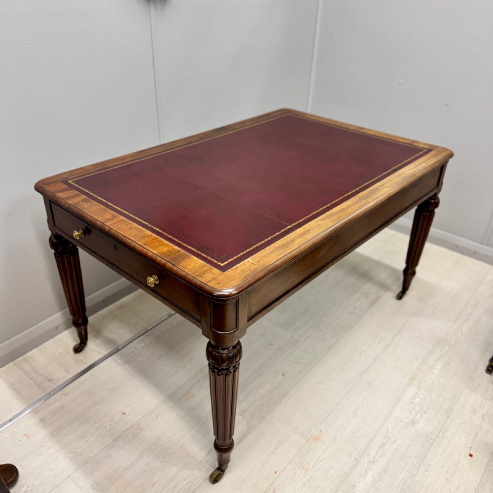 A Regency mahogany library table featuring an inset burgundy leather writing surface, turned fluted legs on brass castors, and a side drawer with brass knobs. View VII
