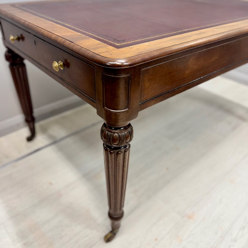 A Regency mahogany library table featuring an inset burgundy leather writing surface, turned fluted legs on brass castors, and a side drawer with brass knobs. View IV