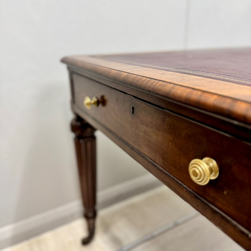 A Regency mahogany library table featuring an inset burgundy leather writing surface, turned fluted legs on brass castors, and a side drawer with brass knobs. View VI