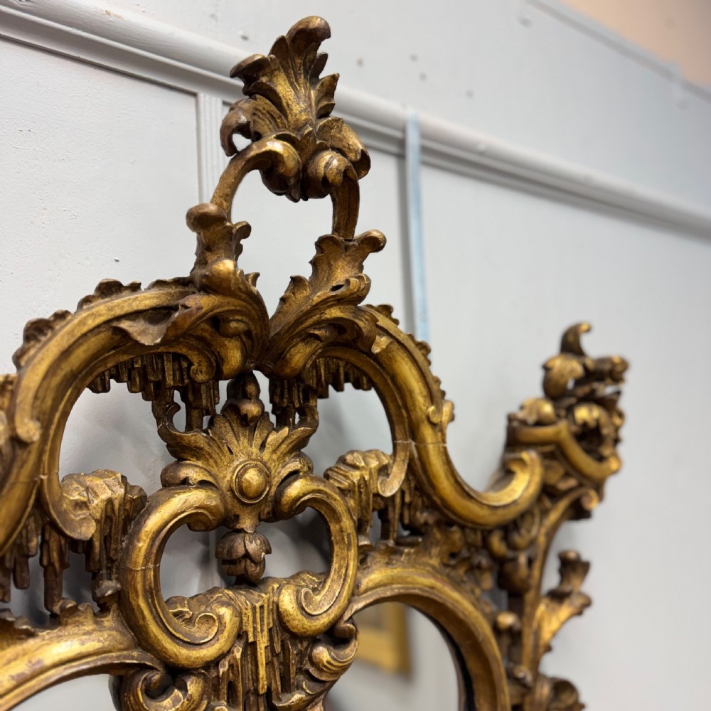 An ornate 18th-century carved giltwood wall mirror featuring elaborate rococo scrolling acanthus leaves and a central arched frame. View IV