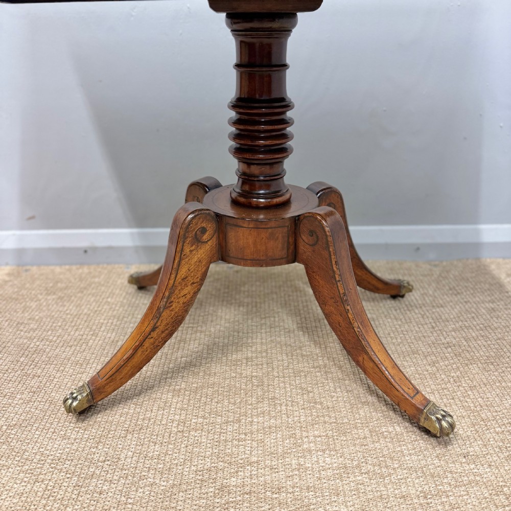 A Georgian satinwood games table featuring ebony line inlay, a turned central pedestal, and four splayed legs with brass paw feet. View IV