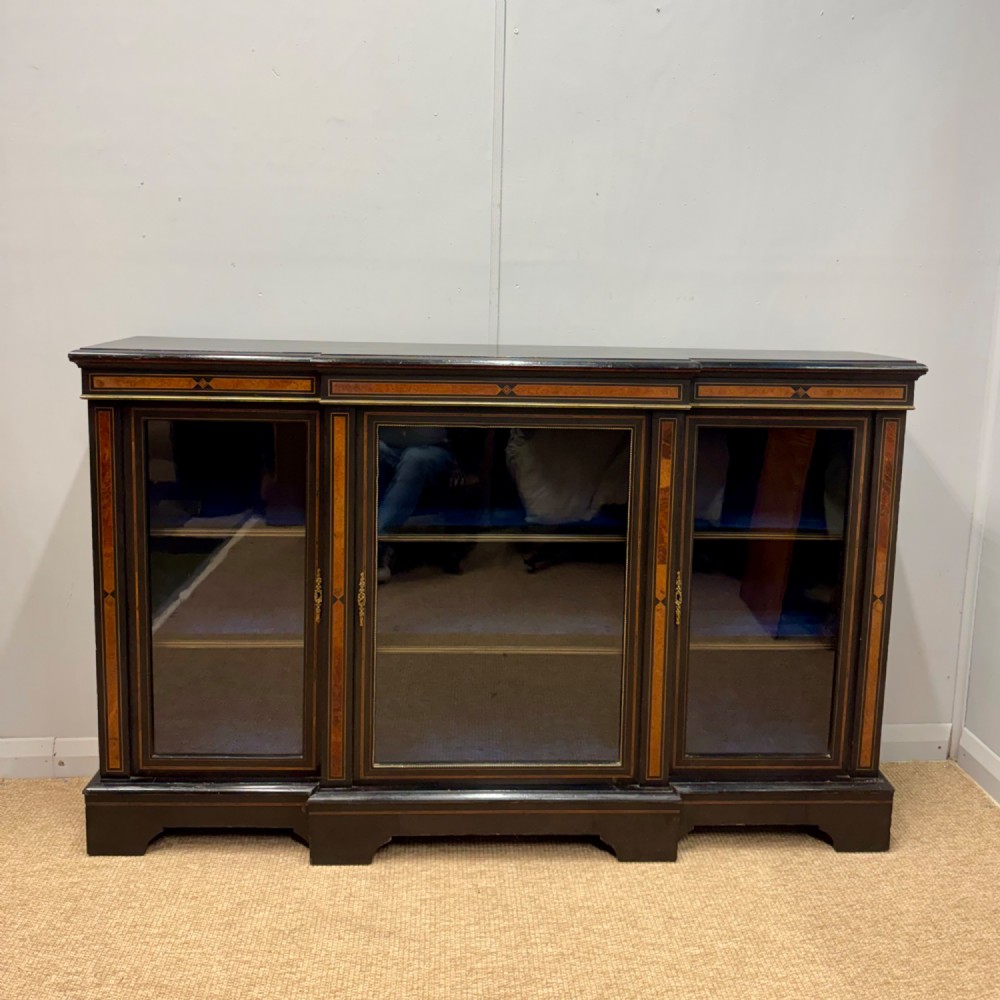 A late 19th-century low ebonised breakfront bookcase featuring three glazed doors, decorative wood inlay along the frieze and pilasters, and brass mounts.