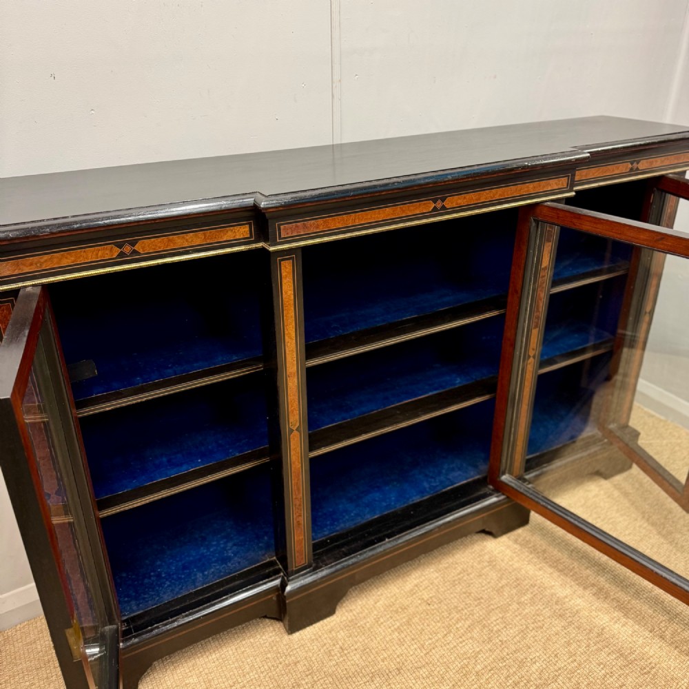 A late 19th-century low ebonised breakfront bookcase featuring three glazed doors, decorative wood inlay along the frieze and pilasters, and brass mounts. View VII