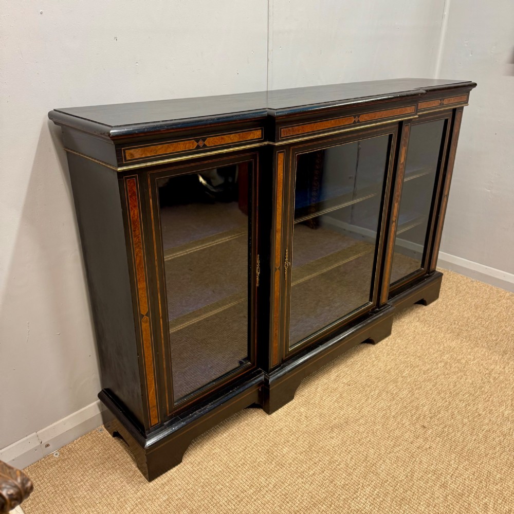 A late 19th-century low ebonised breakfront bookcase featuring three glazed doors, decorative wood inlay along the frieze and pilasters, and brass mounts. View VIII