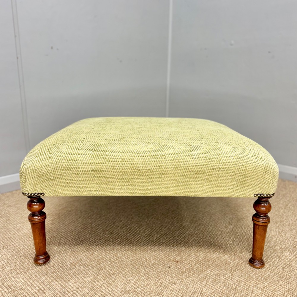 A square footstool upholstered in light green herringbone fabric featuring brass stud trim and four dark turned wooden legs.