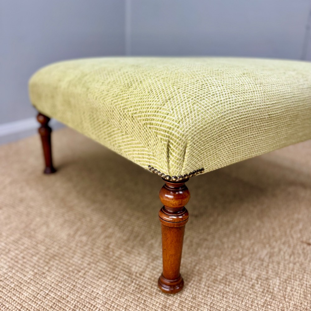 A square footstool upholstered in light green herringbone fabric featuring brass stud trim and four dark turned wooden legs. View II