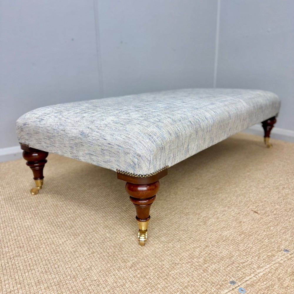 A low rectangular upholstered footstool featuring light blue and grey woven fabric, decorative brass tack trim, and turned dark mahogany legs on brass castors.