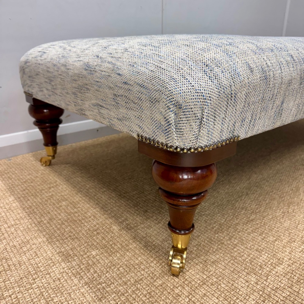A low rectangular upholstered footstool featuring light blue and grey woven fabric, decorative brass tack trim, and turned dark mahogany legs on brass castors. View II