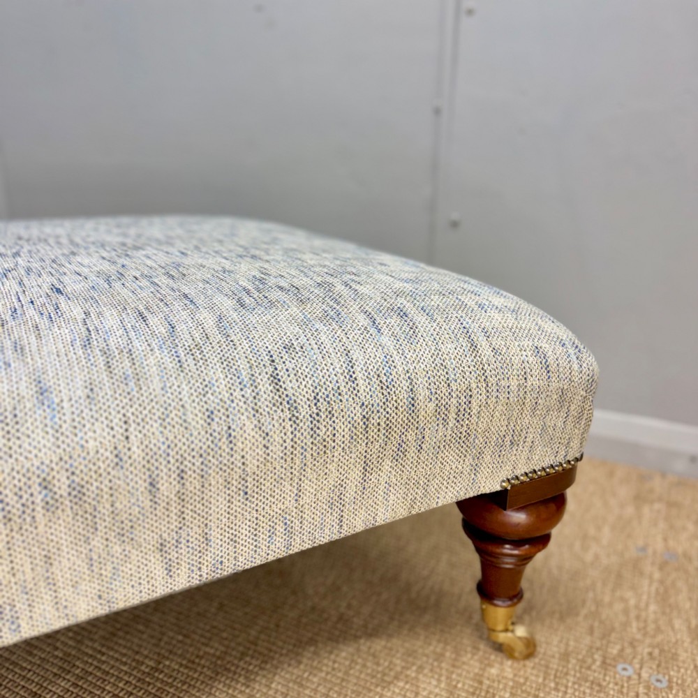 A low rectangular upholstered footstool featuring light blue and grey woven fabric, decorative brass tack trim, and turned dark mahogany legs on brass castors. View III