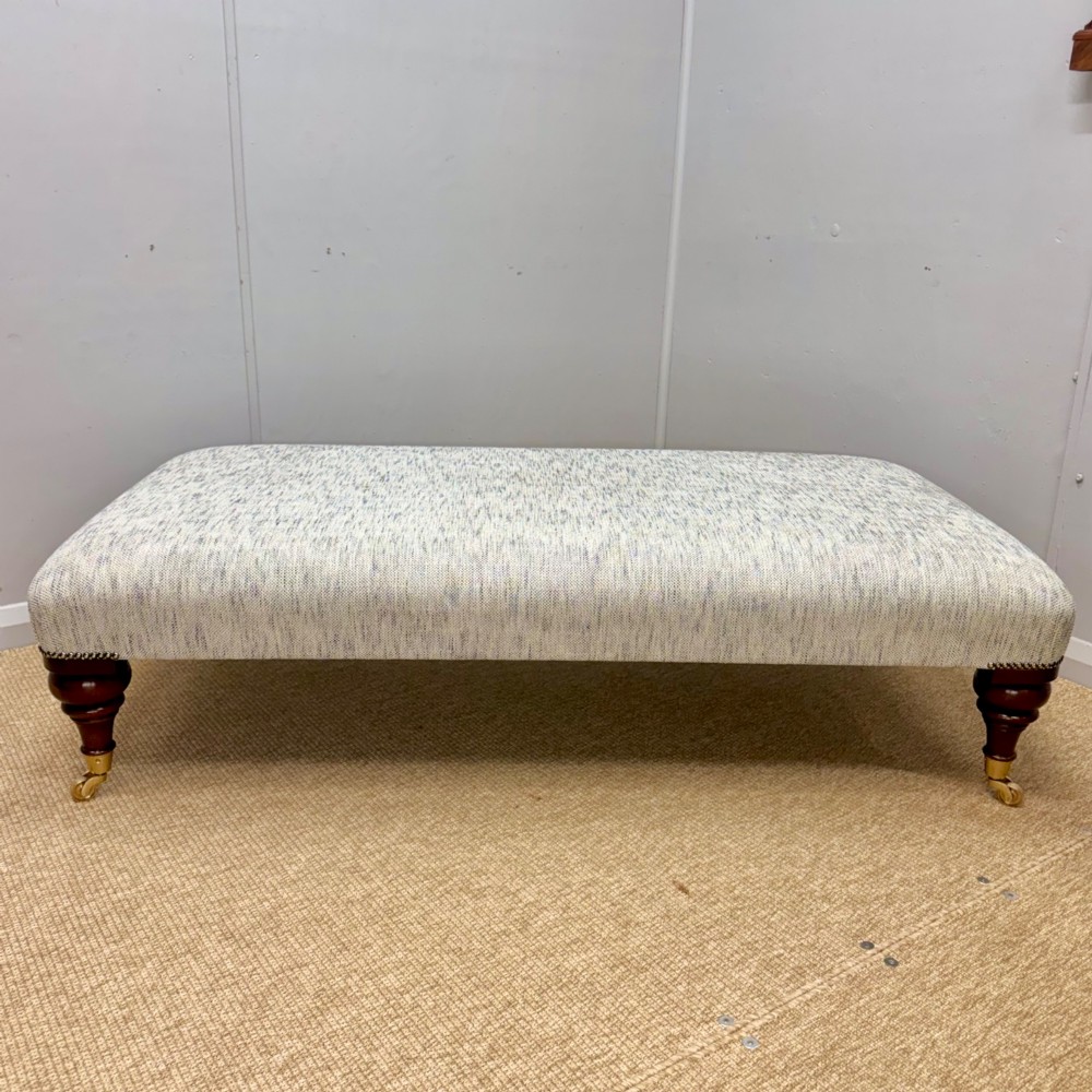 A low rectangular upholstered footstool featuring light blue and grey woven fabric, decorative brass tack trim, and turned dark mahogany legs on brass castors. View IV