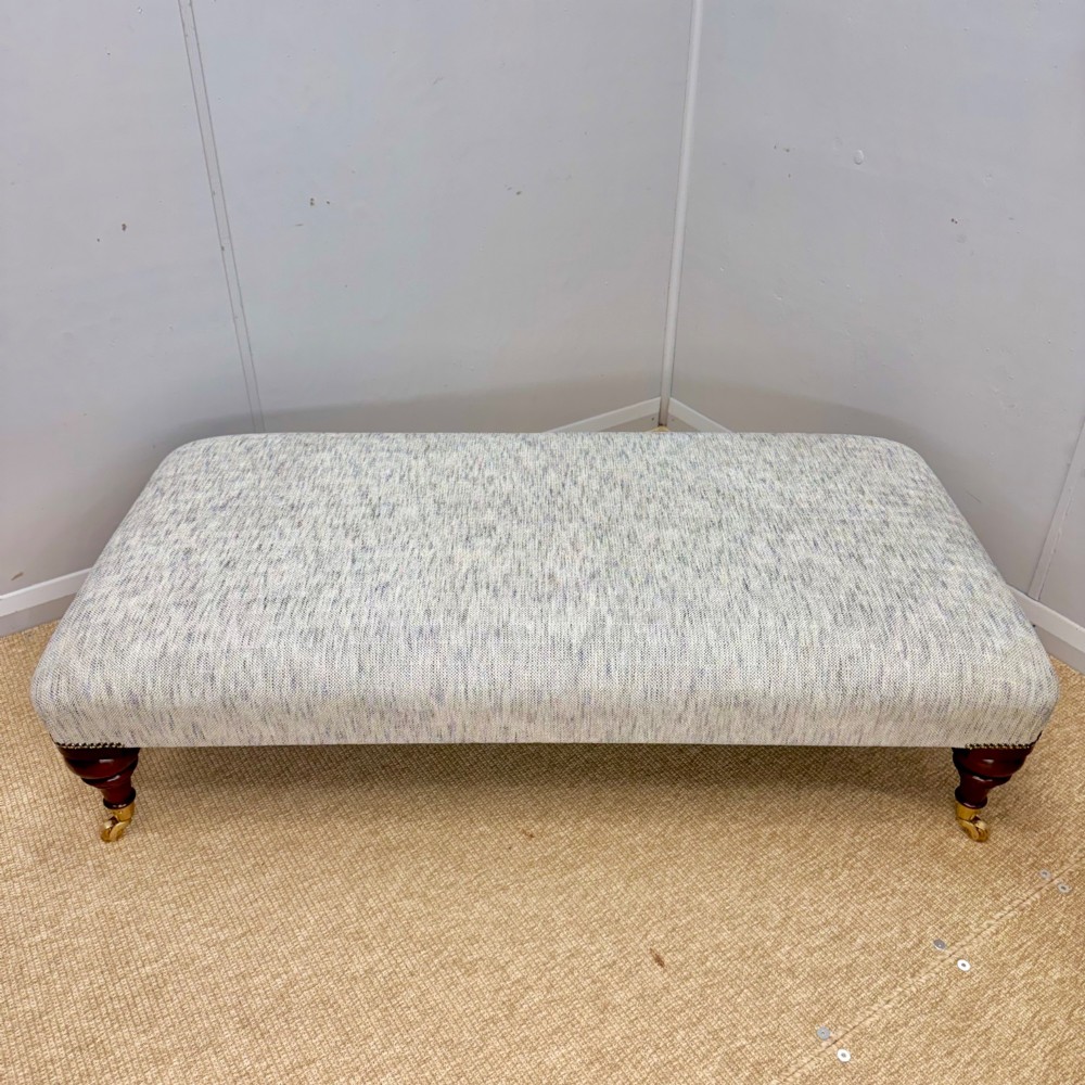 A low rectangular upholstered footstool featuring light blue and grey woven fabric, decorative brass tack trim, and turned dark mahogany legs on brass castors. View V