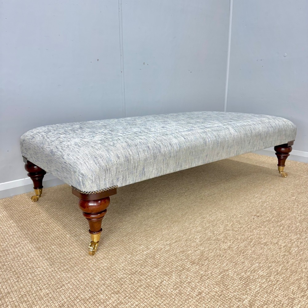 A low rectangular upholstered footstool featuring light blue and grey woven fabric, decorative brass tack trim, and turned dark mahogany legs on brass castors. View VI