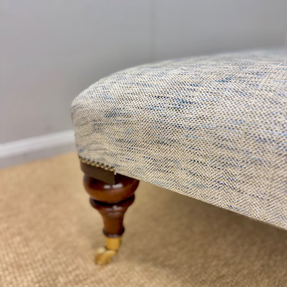 A low rectangular upholstered footstool featuring light blue and grey woven fabric, decorative brass tack trim, and turned dark mahogany legs on brass castors. View VII