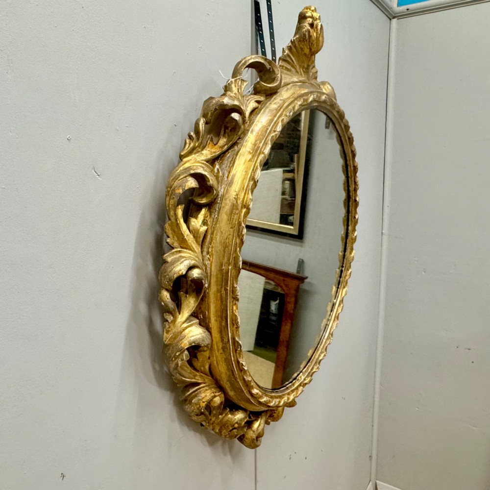 A circular giltwood wall mirror framed in carved acanthus leaves and topped with a shell crest. View IV