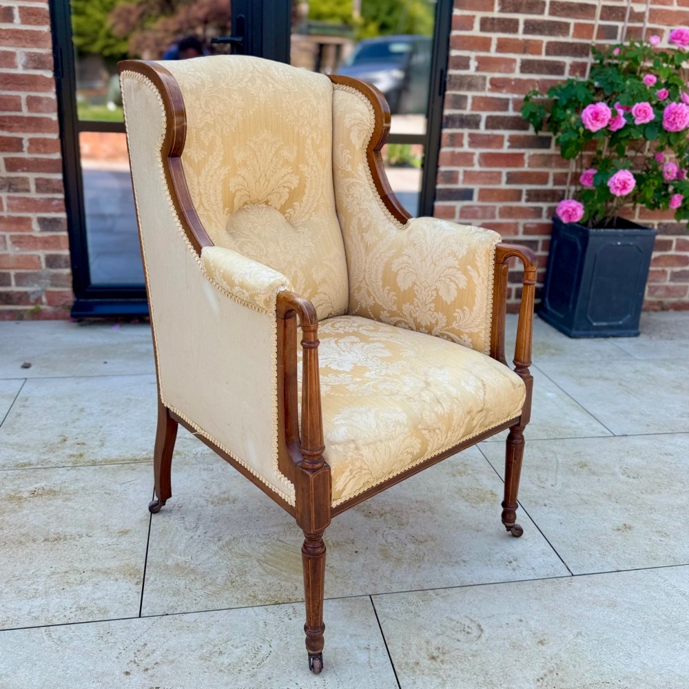 An Edwardian inlaid mahogany wing armchair upholstered in gold patterned damask with turned front legs on brass castors.