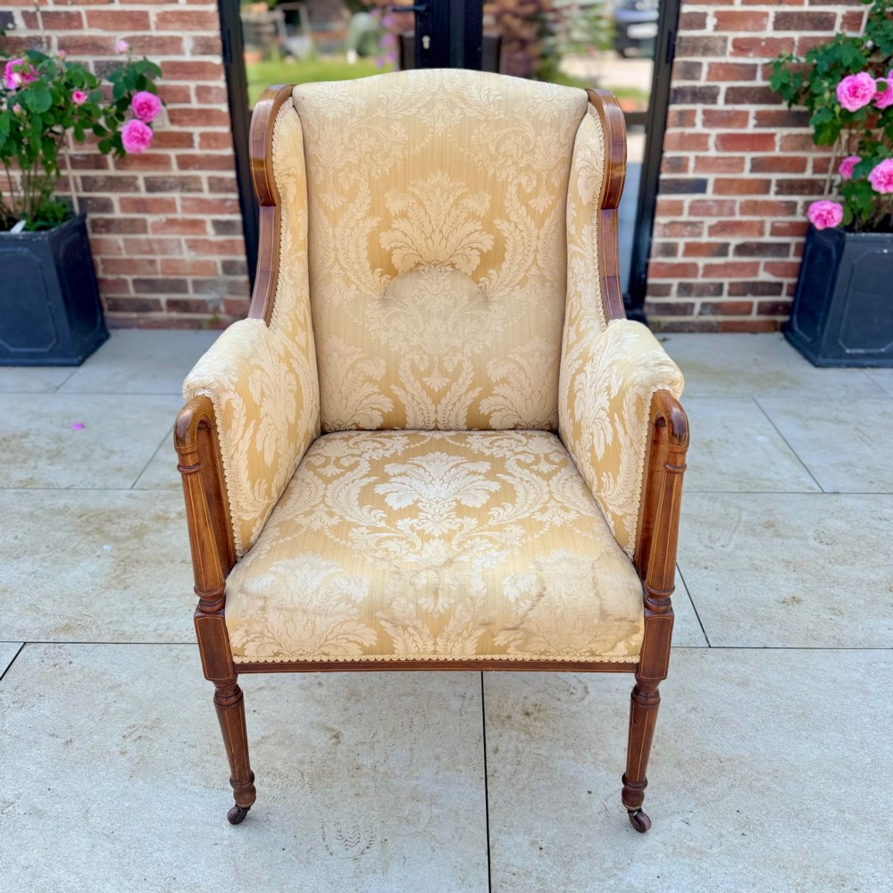 An Edwardian inlaid mahogany wing armchair upholstered in gold patterned damask with turned front legs on brass castors. View II