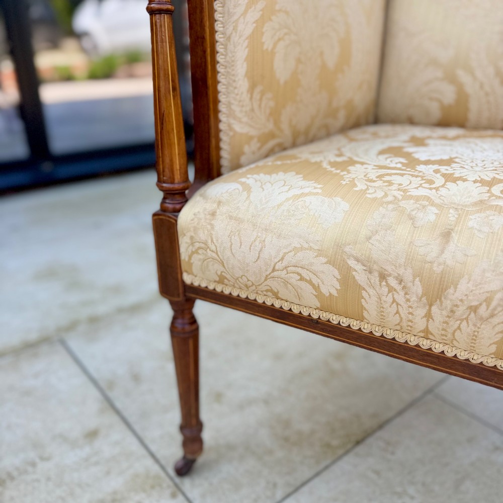 An Edwardian inlaid mahogany wing armchair upholstered in gold patterned damask with turned front legs on brass castors. View VI