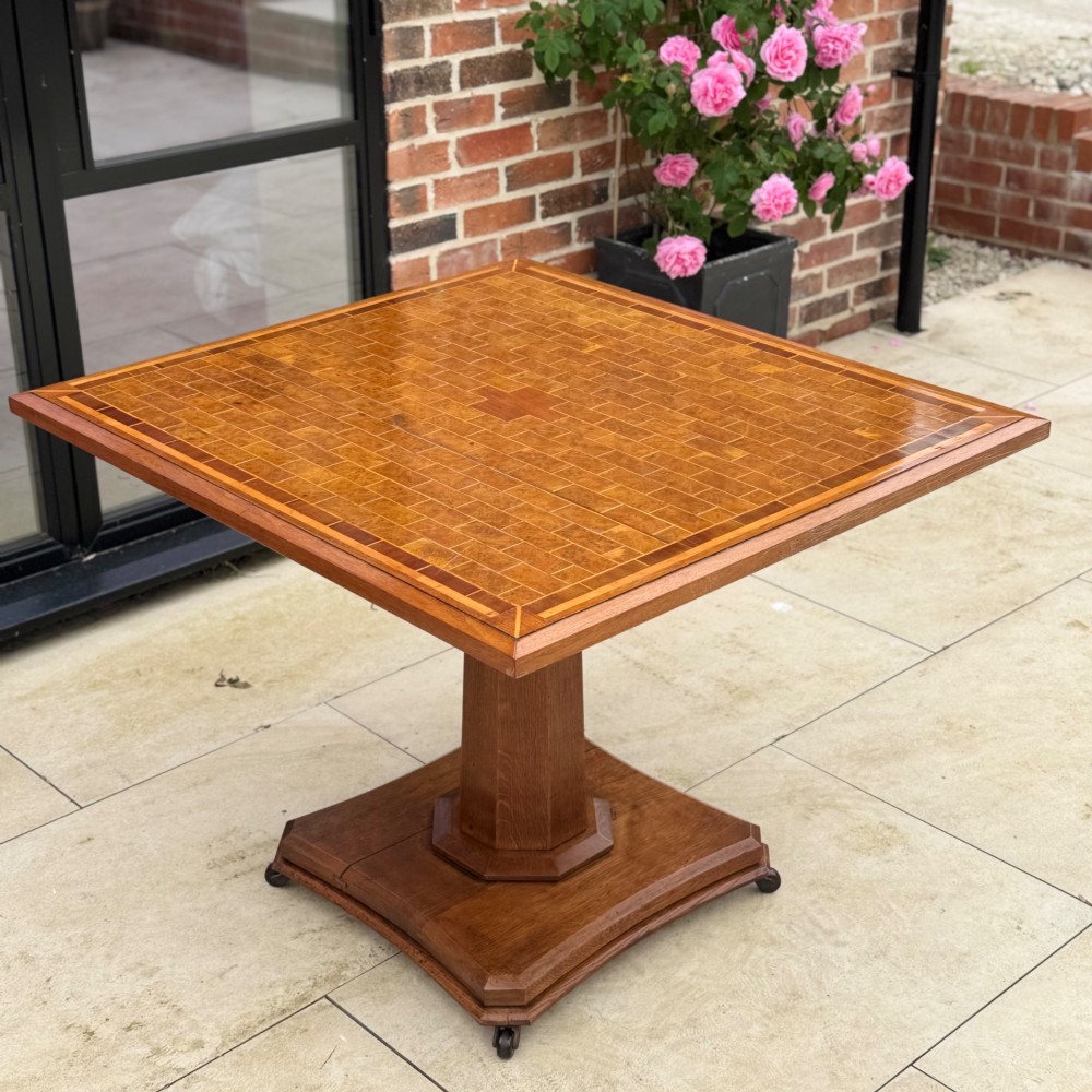 A square antique tilt-top centre table featuring a geometric burr walnut parquetry top framed by an oak border, supported on a faceted pedestal base with castors.