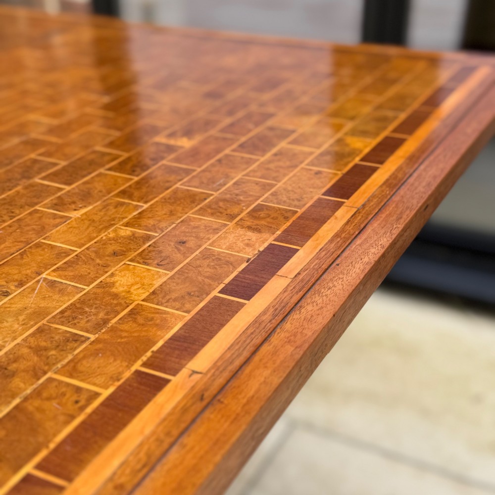 A square antique tilt-top centre table featuring a geometric burr walnut parquetry top framed by an oak border, supported on a faceted pedestal base with castors. View II