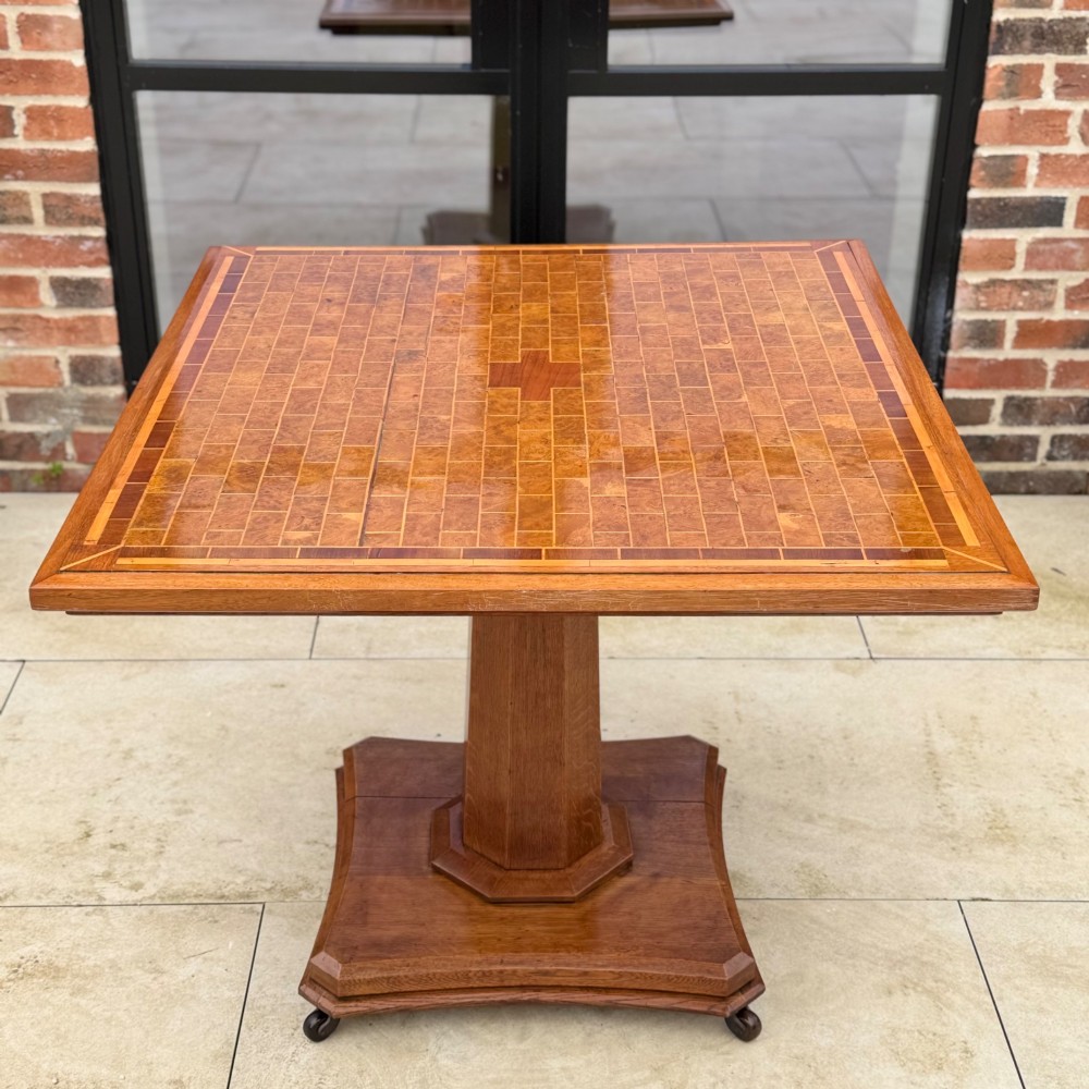 A square antique tilt-top centre table featuring a geometric burr walnut parquetry top framed by an oak border, supported on a faceted pedestal base with castors. View III