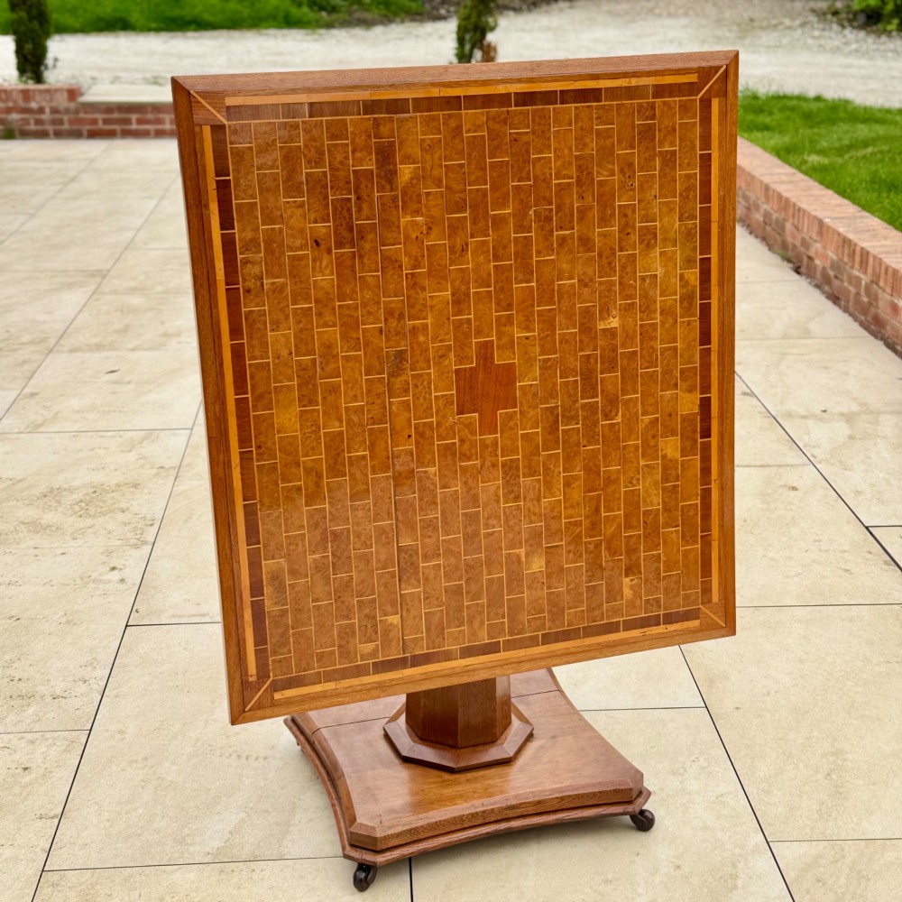 A square antique tilt-top centre table featuring a geometric burr walnut parquetry top framed by an oak border, supported on a faceted pedestal base with castors. View IV
