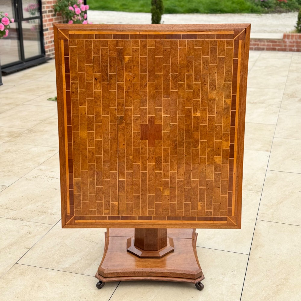 A square antique tilt-top centre table featuring a geometric burr walnut parquetry top framed by an oak border, supported on a faceted pedestal base with castors. View V