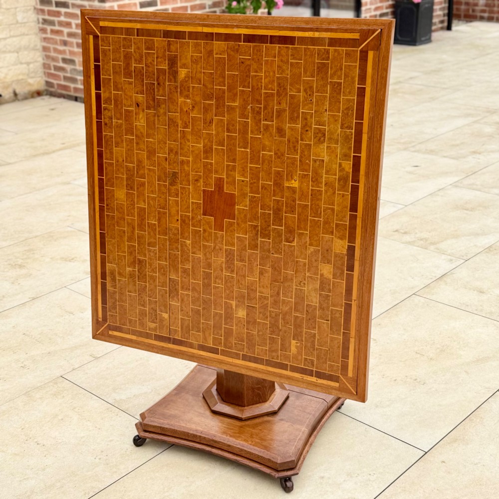 A square antique tilt-top centre table featuring a geometric burr walnut parquetry top framed by an oak border, supported on a faceted pedestal base with castors. View VI