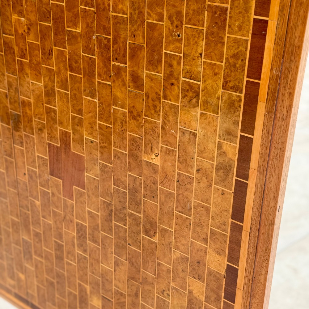 A square antique tilt-top centre table featuring a geometric burr walnut parquetry top framed by an oak border, supported on a faceted pedestal base with castors. View VII