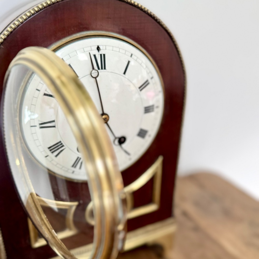 An arched antique French mahogany mantel clock featuring a white face with Roman numerals, decorative brass trim, and a stepped brass base. View V