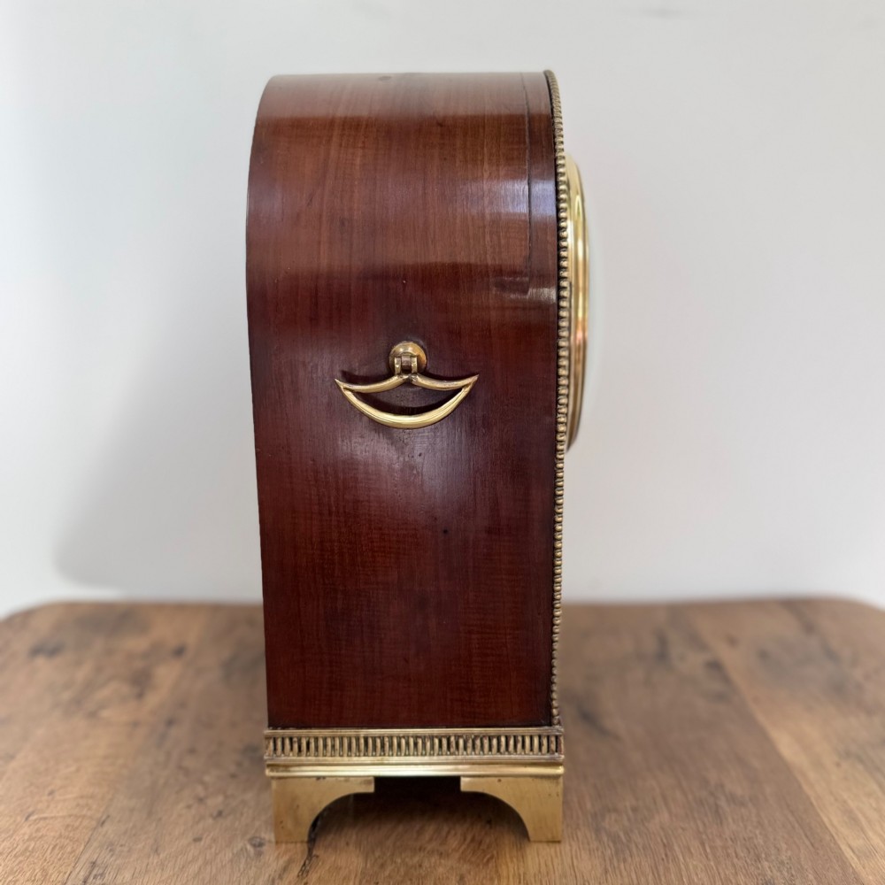 An arched antique French mahogany mantel clock featuring a white face with Roman numerals, decorative brass trim, and a stepped brass base. View VI