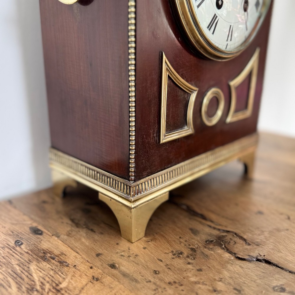 An arched antique French mahogany mantel clock featuring a white face with Roman numerals, decorative brass trim, and a stepped brass base. View VII