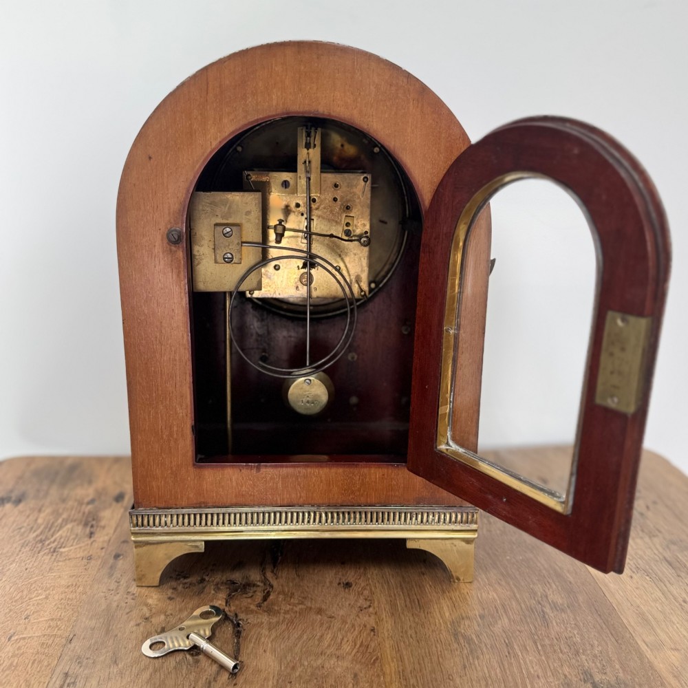 An arched antique French mahogany mantel clock featuring a white face with Roman numerals, decorative brass trim, and a stepped brass base. View VIII