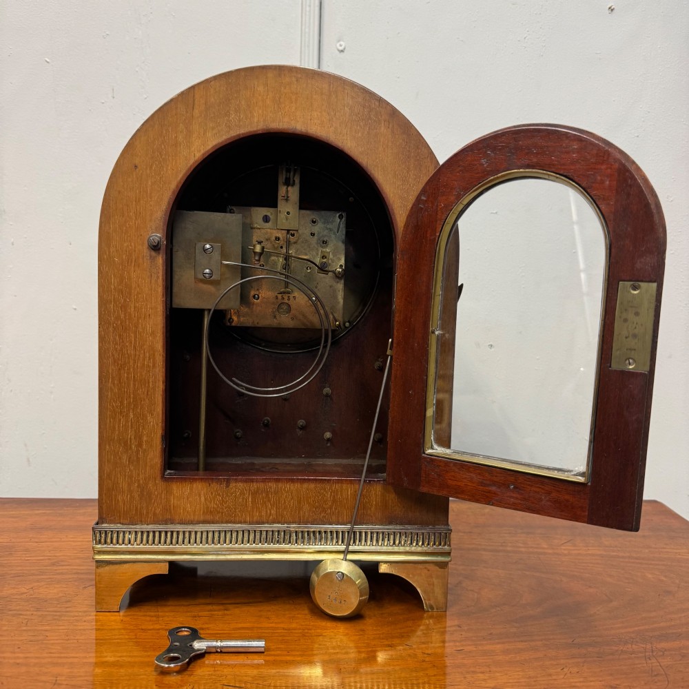 An arched antique French mahogany mantel clock featuring a white face with Roman numerals, decorative brass trim, and a stepped brass base. View IX