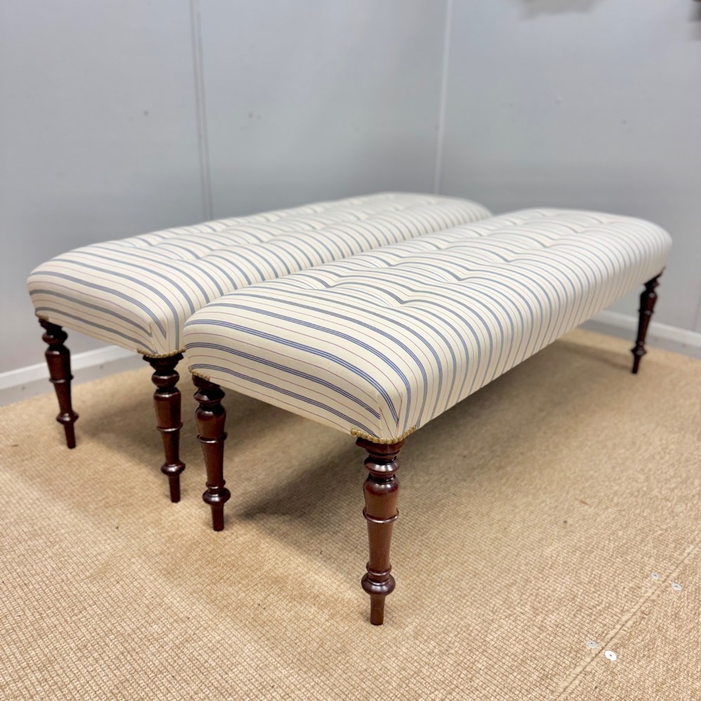 A pair of five-foot long upholstered stools with cream and blue striped buttoned ticking fabric set upon dark turned mahogany legs with brass studs.