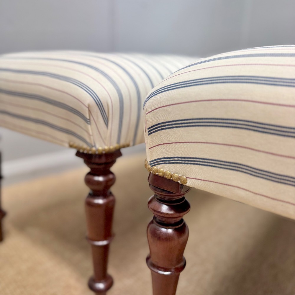 A pair of five-foot long upholstered stools with cream and blue striped buttoned ticking fabric set upon dark turned mahogany legs with brass studs. View IV