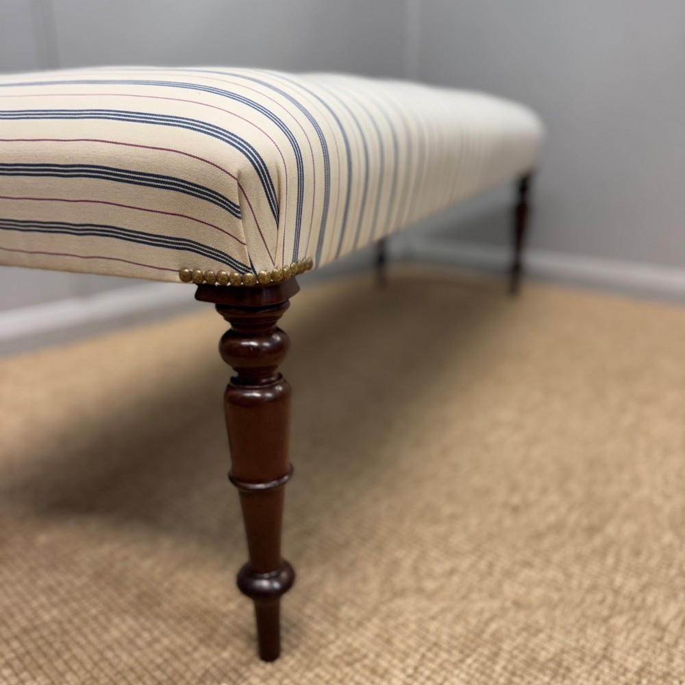 A pair of five-foot long upholstered stools with cream and blue striped buttoned ticking fabric set upon dark turned mahogany legs with brass studs. View V