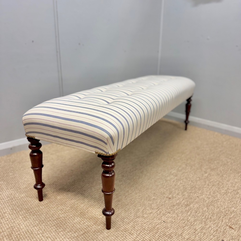 A pair of five-foot long upholstered stools with cream and blue striped buttoned ticking fabric set upon dark turned mahogany legs with brass studs. View VI