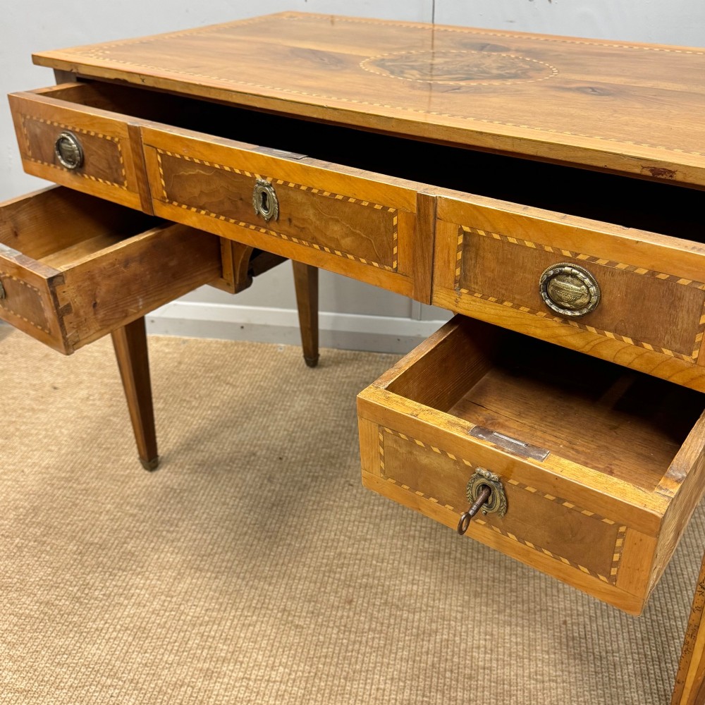 An early nineteenth-century freestanding wood writing desk featuring intricate marquetry inlays, five drawers with brass handles, an arched kneehole, and square tapered legs. View V