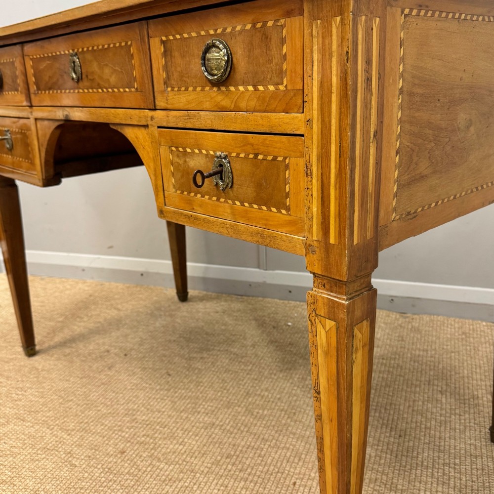 An early nineteenth-century freestanding wood writing desk featuring intricate marquetry inlays, five drawers with brass handles, an arched kneehole, and square tapered legs. View VII
