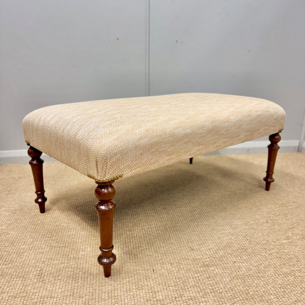 An oblong footstool upholstered in a neutral textured cream fabric supported by four turned warm brown timber legs.