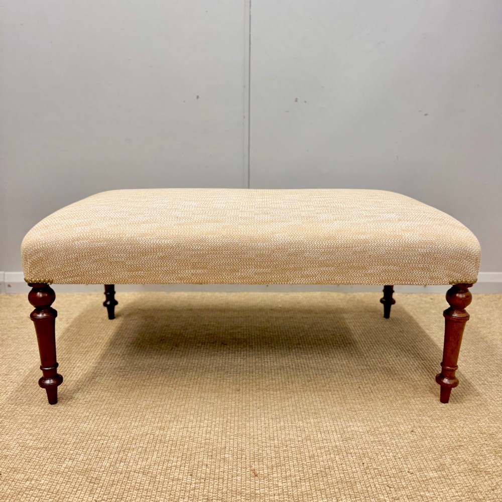 An oblong footstool upholstered in a neutral textured cream fabric supported by four turned warm brown timber legs. View II